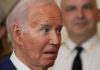Close-up of a man with a surprised expression, wearing a suit, with another figure blurred in the background