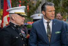 A military officer in uniform stands next to a man in a suit during a news broadcast